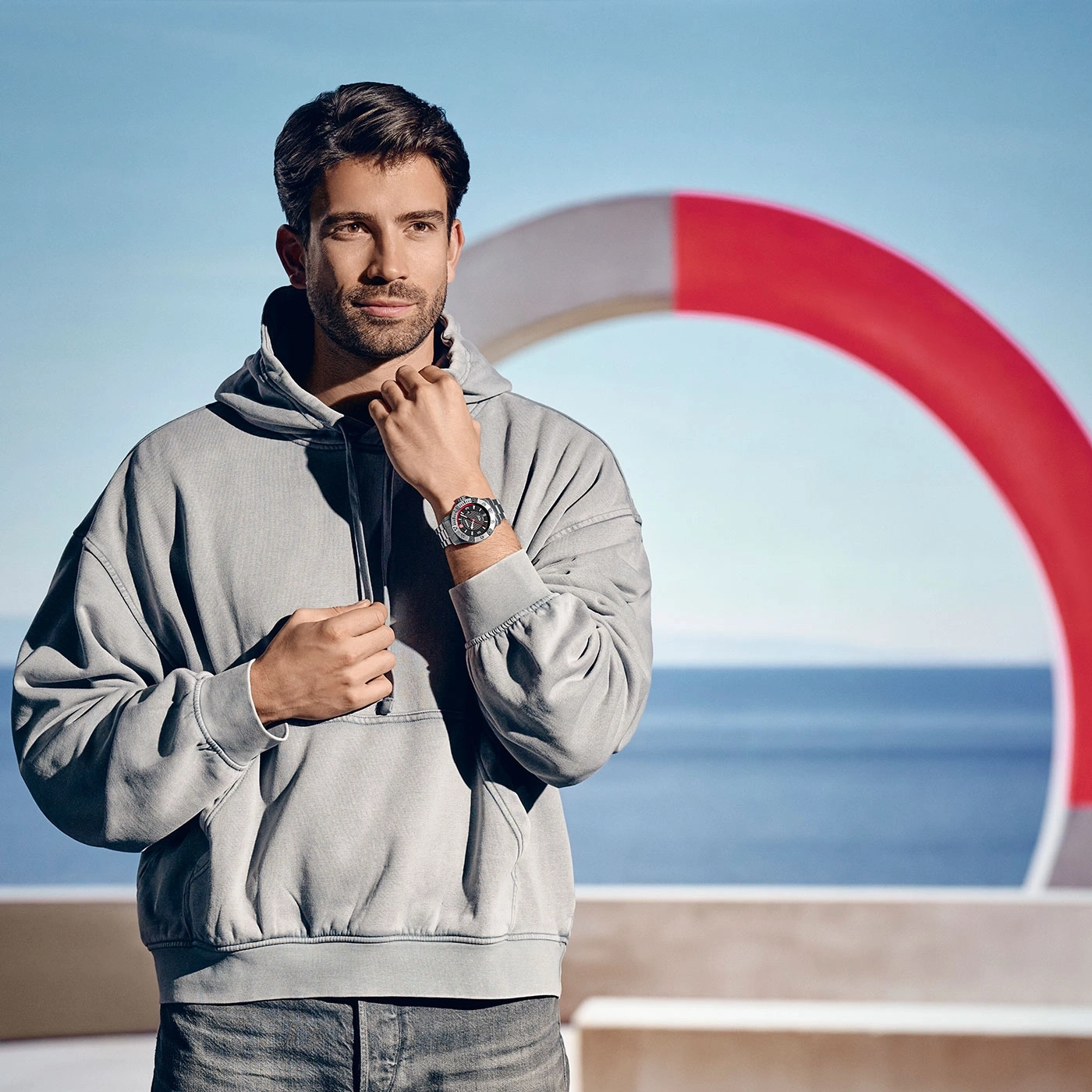 Man wearing watch and hoodie in front of sea and lifebuoy