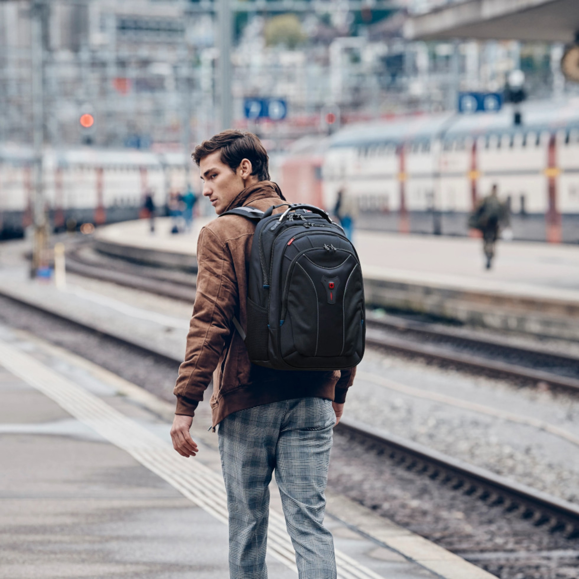 Man with backpack at train station, looking back, brown jacket, plaid pants, train