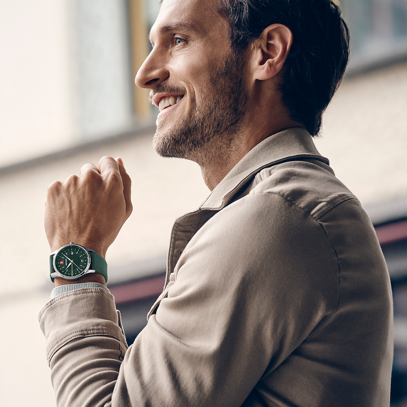 A man wearing a green watch and beige jacket smiles.