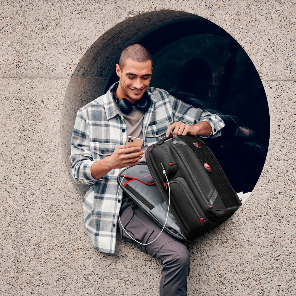 Man sitting in opening, looking at phone, backpack, headphones, smiling, outdoors