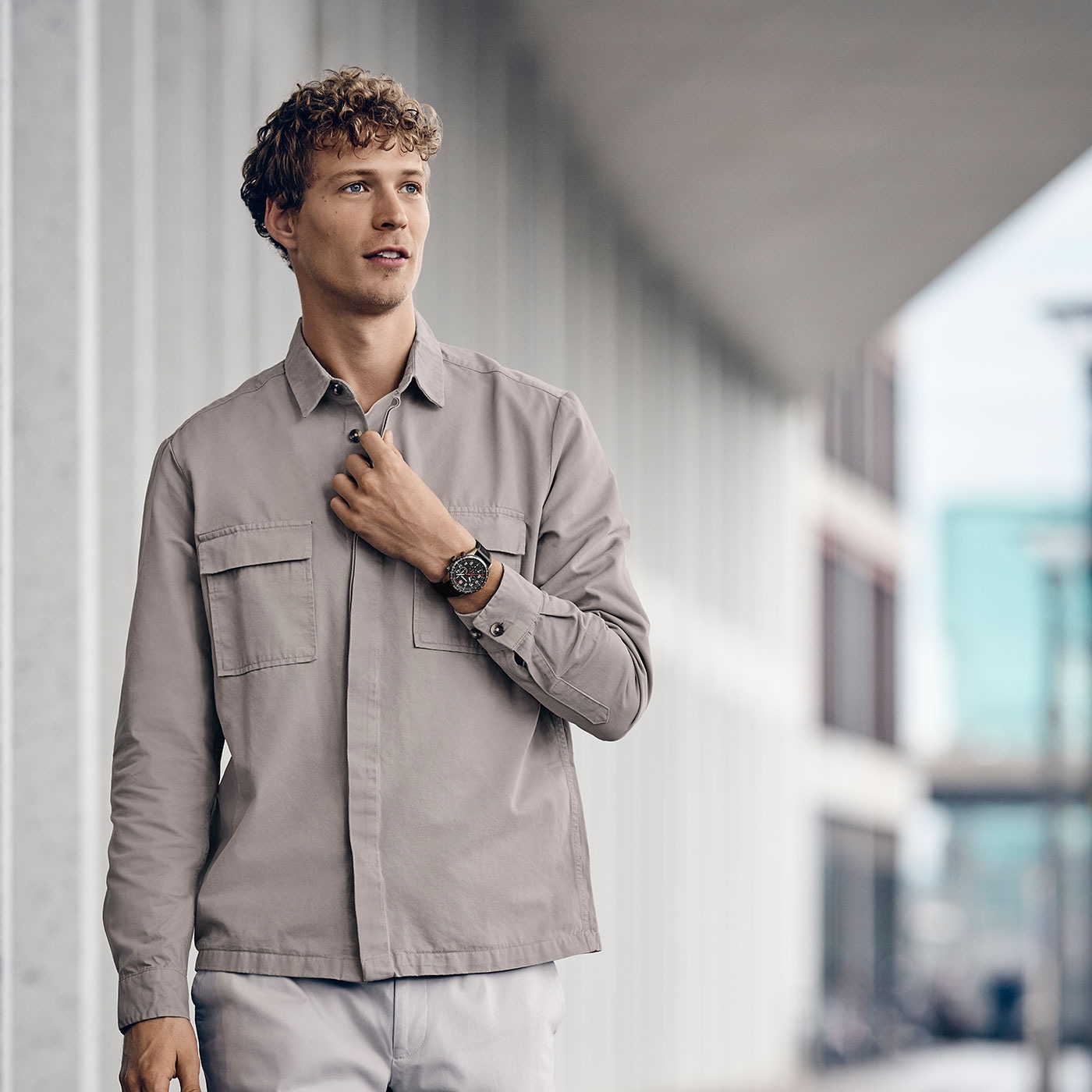 Man wearing a watch, buttoning a shirt in a modern urban environment.
