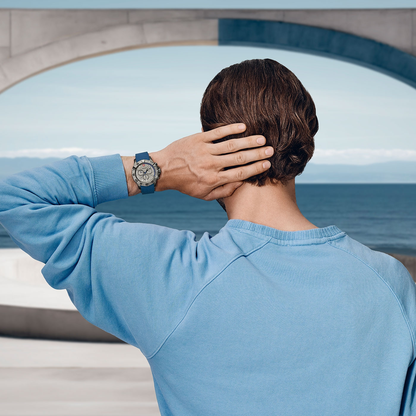 Man with watch relaxing by the sea, blue sweater, looking at the water, hand on head.
