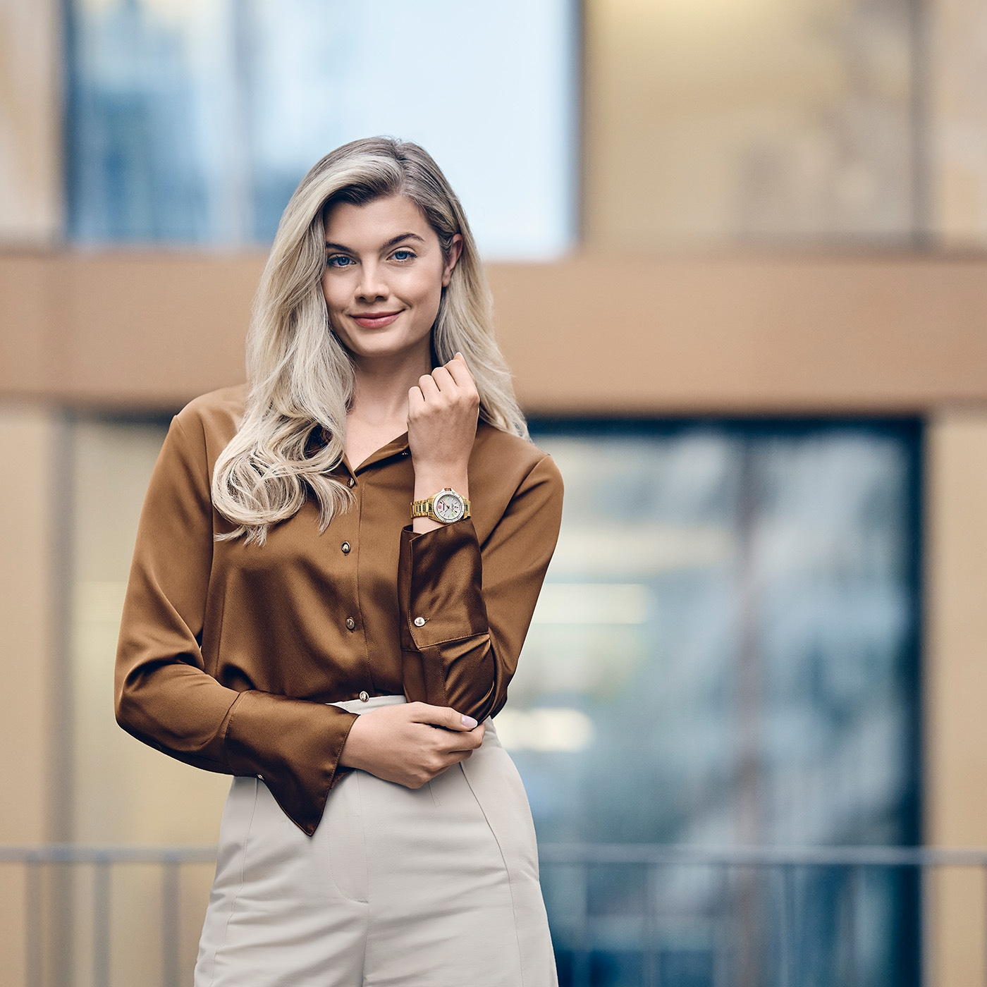 Woman wearing gold watch, brown blouse, and cream pants outdoors.