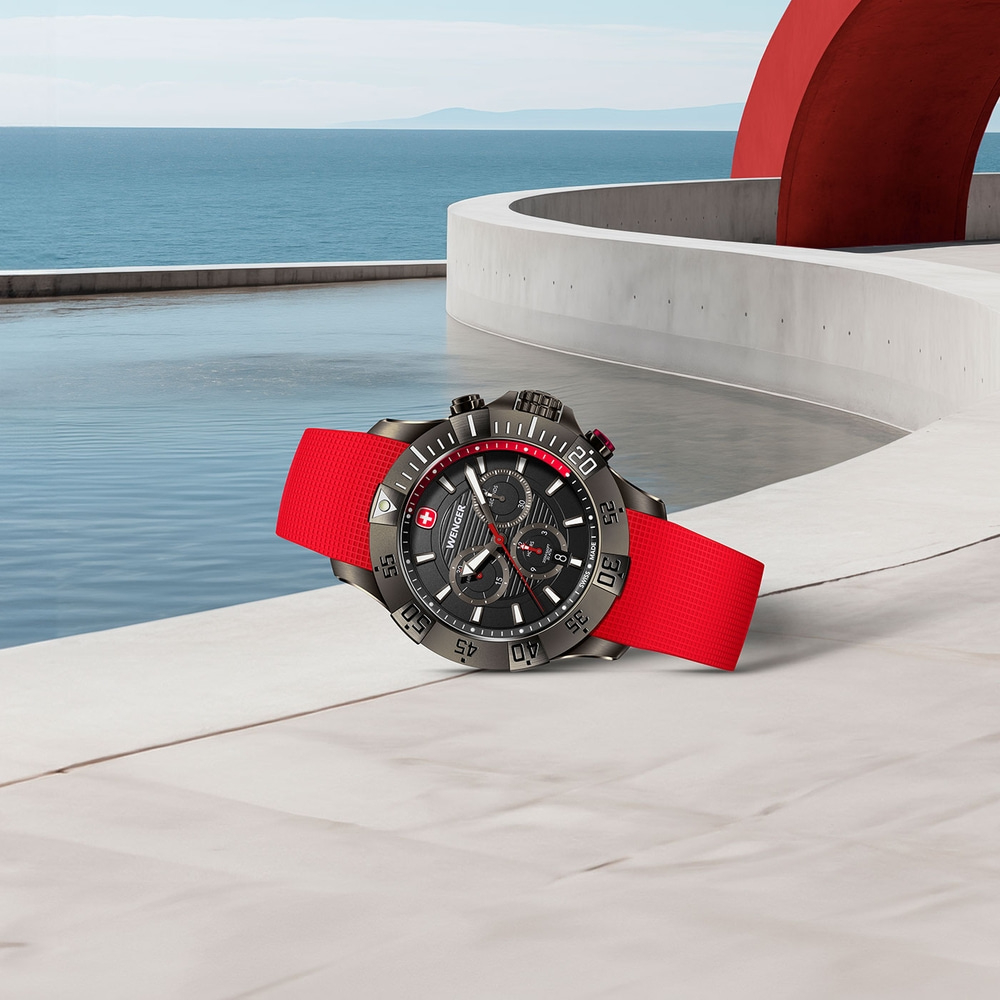 Watch with red strap on white surface with ocean view and red sculpture in the background.