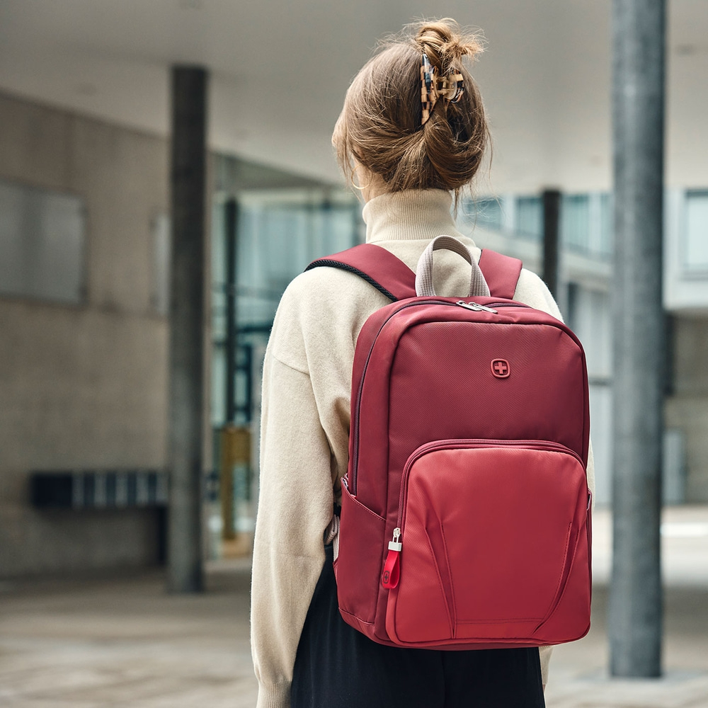Person with a red backpack in front of a building, back view, hair up.