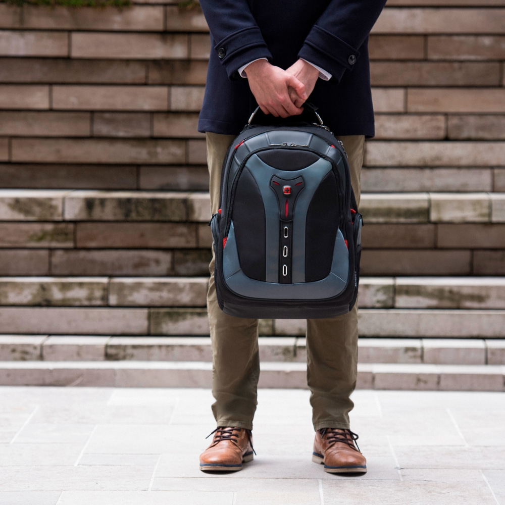 Man holding backpack in front of stairs. He wears jacket, pants, and shoes.