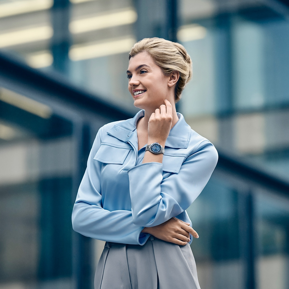 Woman in light blue blouse and gray skirt looking at a watch.