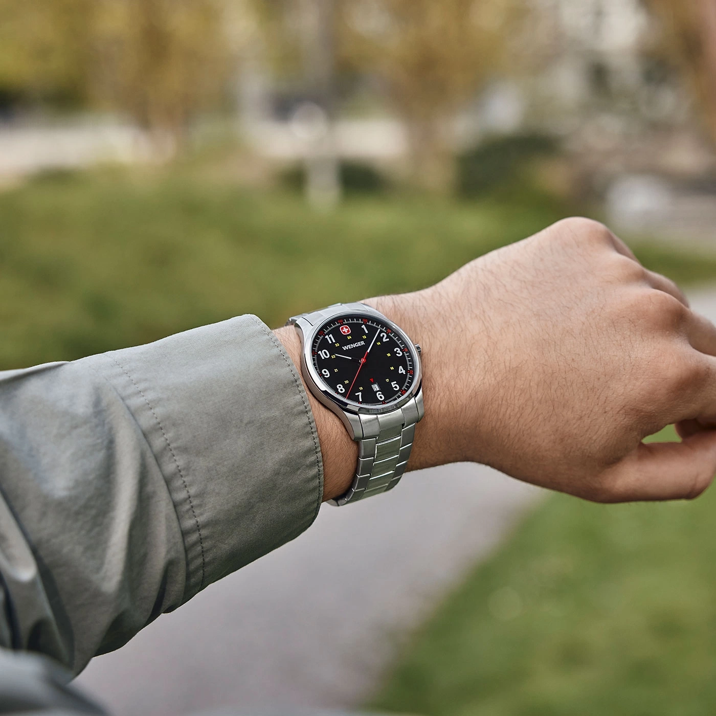 Watch on wrist, silver bracelet, black dial, Wenger logo, outdoors, blurred background