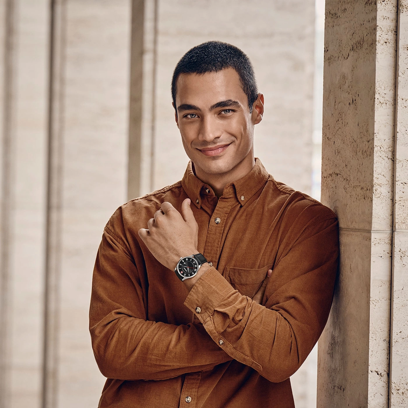 Man wearing a watch, leaning against a column, smiling at the camera.