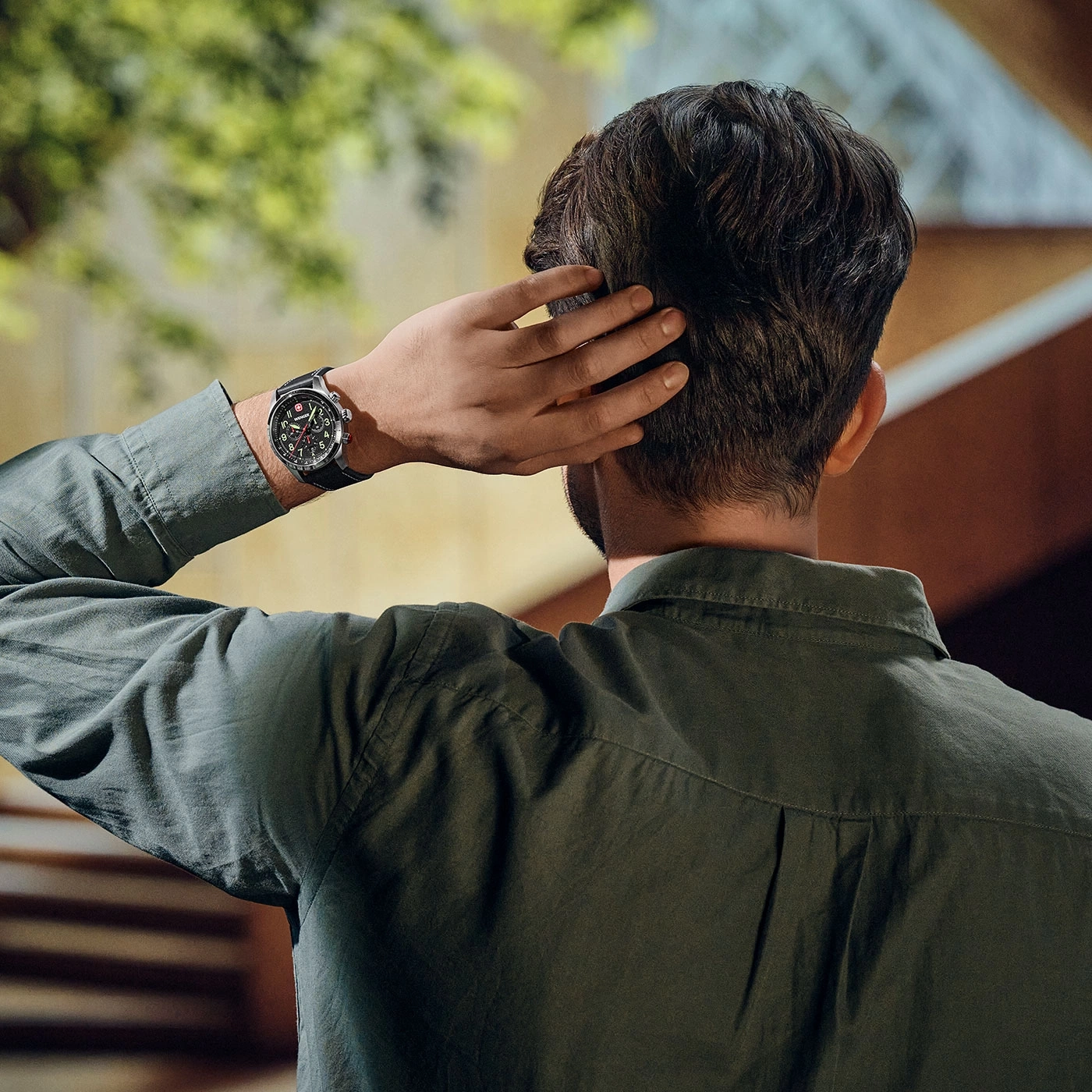Man with watch scratching back of head, wearing a green shirt, outdoors.