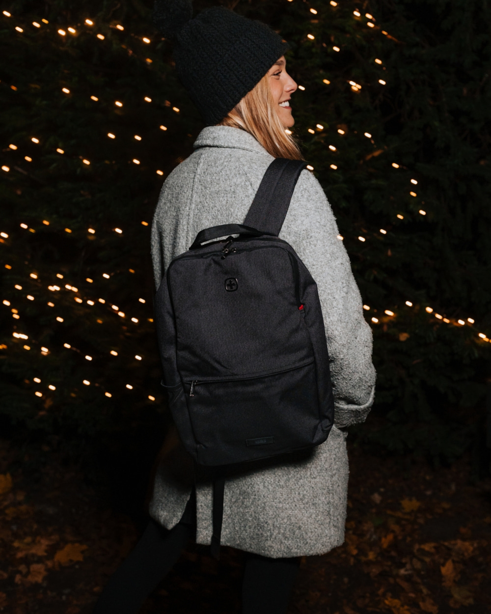 Woman with backpack and hat smiles in front of Christmas lights at night
