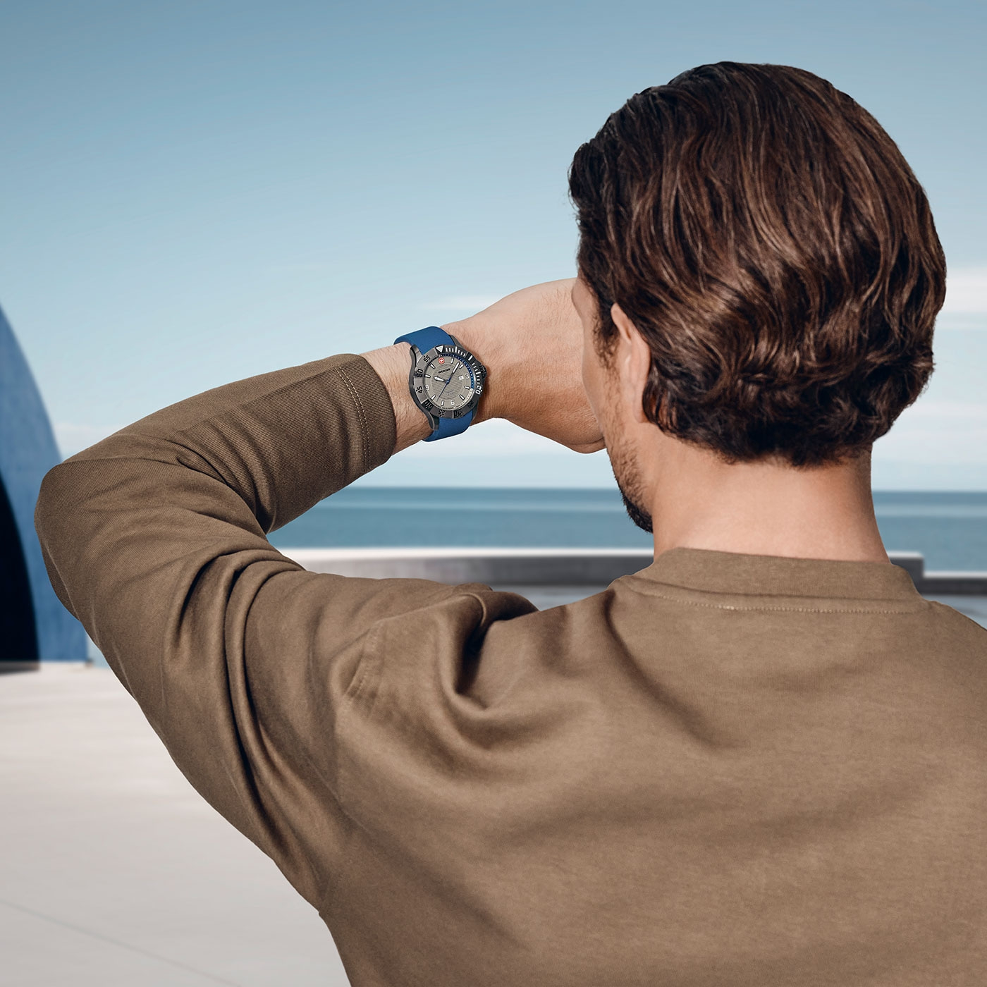 Man with watch looking at the sea, brown shirt, blue background, back view