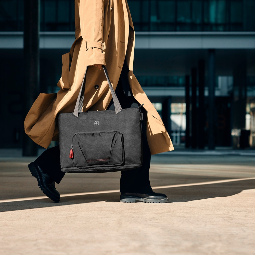 Person carrying black bag and beige coat in front of a building.