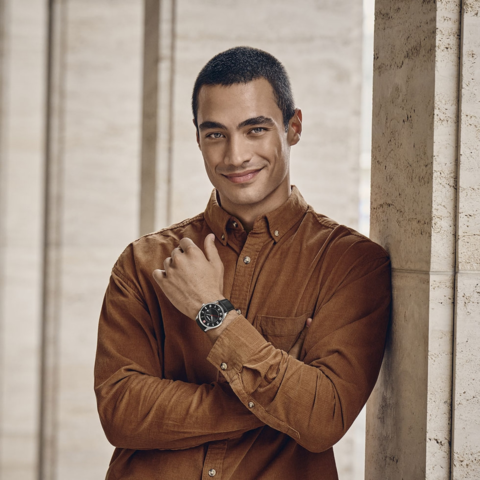 Man wearing watch, leaning against column, smiling, brown shirt, portrait.