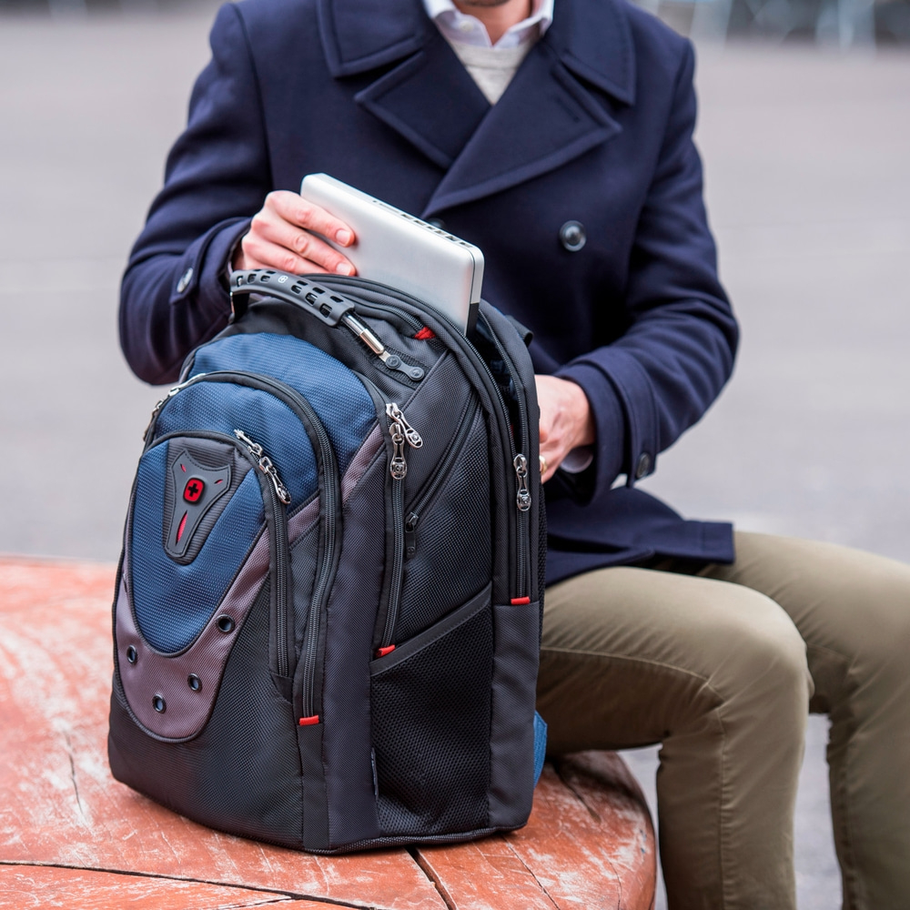 Man putting laptop in backpack. Person sitting on bench. Blue jacket and pants.