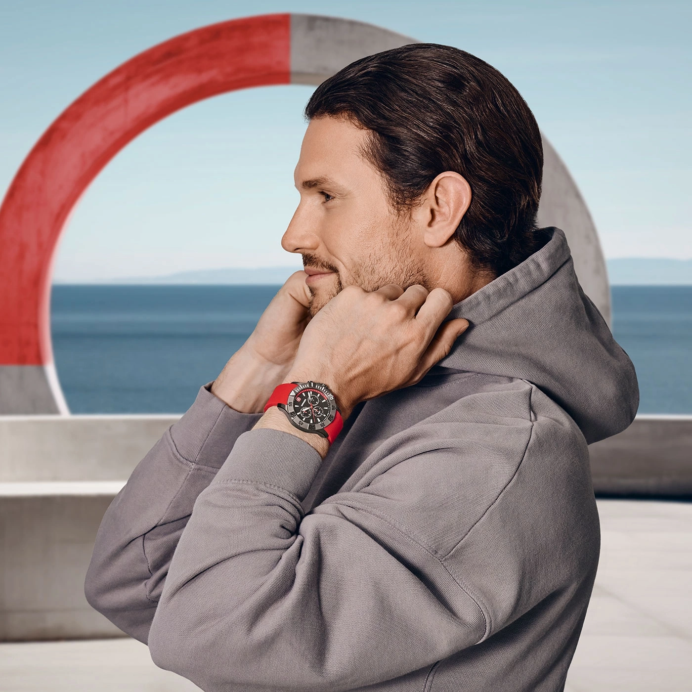 Mann mit Uhr und rotem Armband, vor einem roten Ring, Meer im Hintergrund