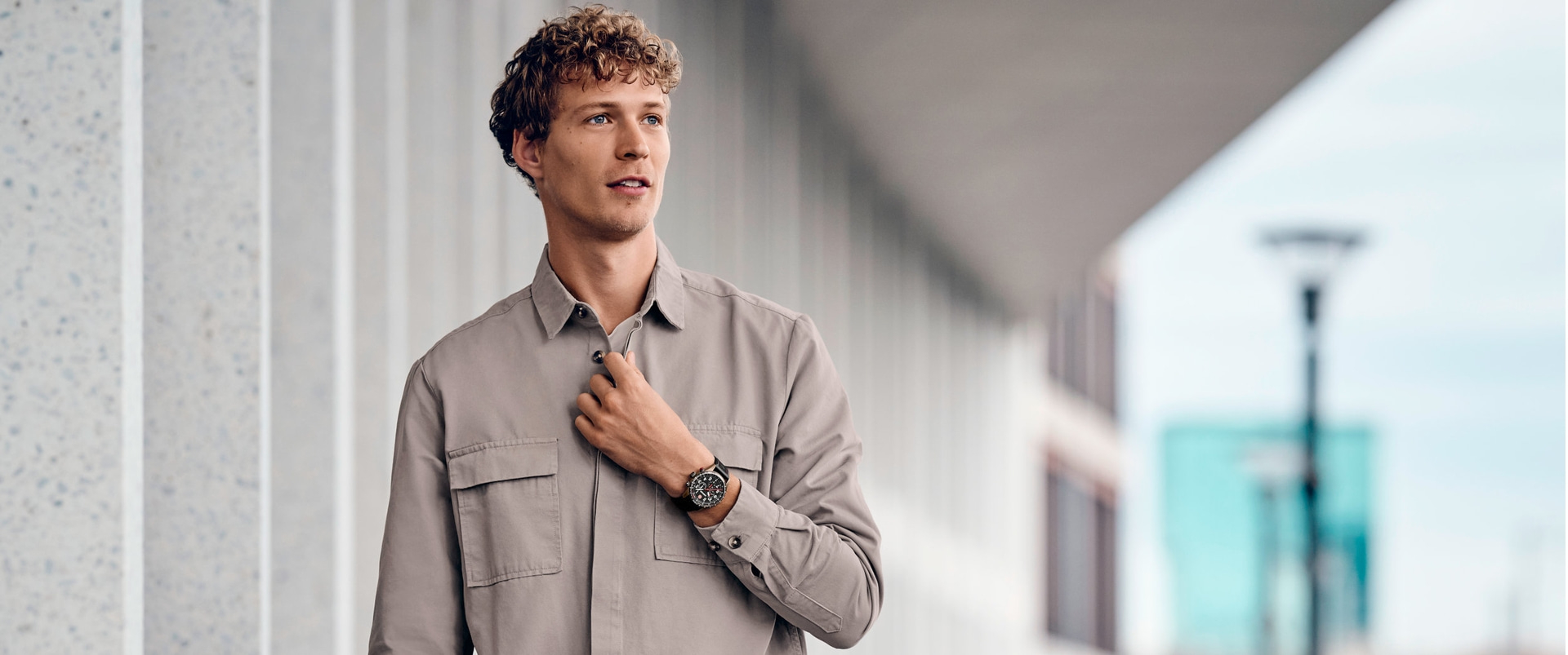 A man in a shirt wearing a watch standing by a building, looking away.