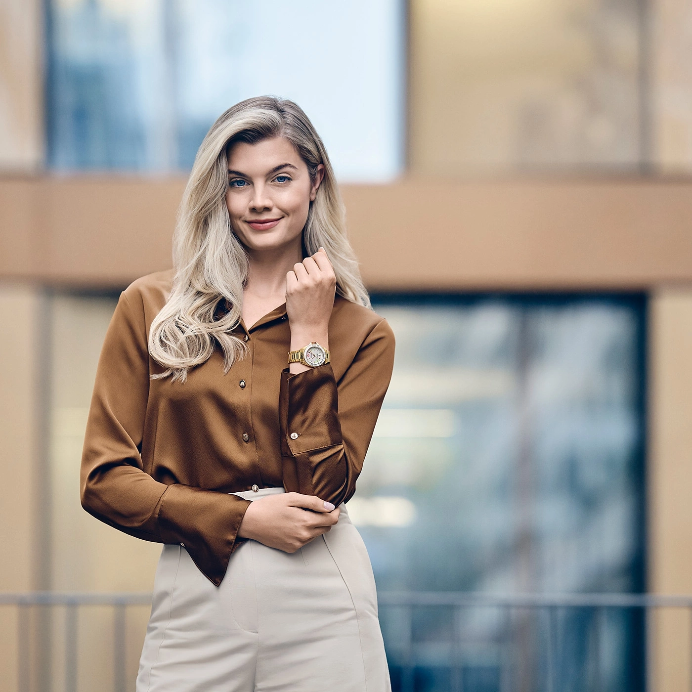Woman wearing gold watch, brown blouse, and cream pants outdoors.