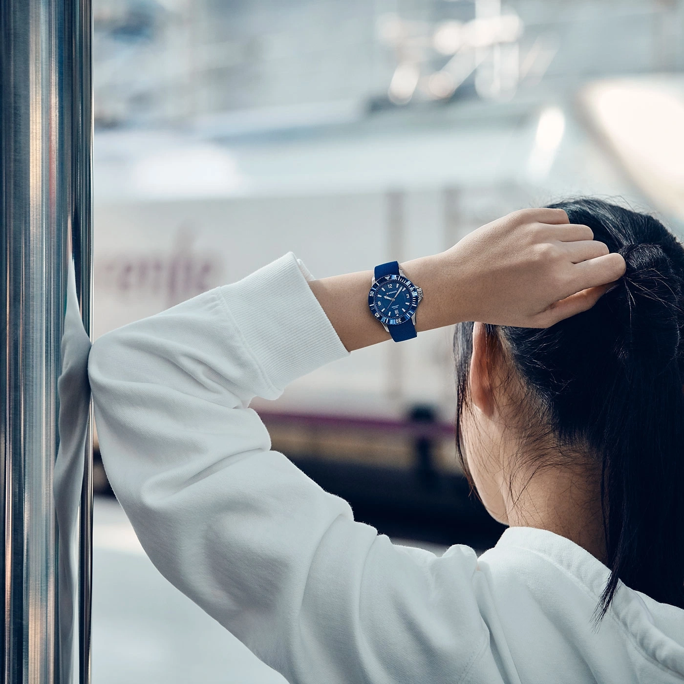 Woman with watch looking out window, wearing a white hoodie, train in background.