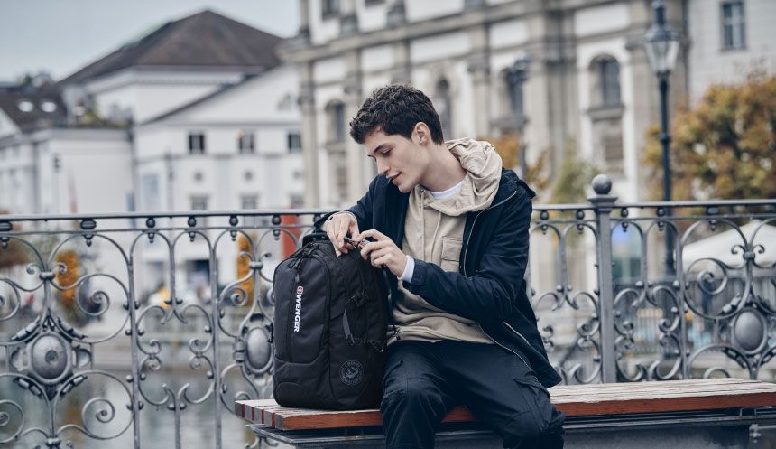 Young man sitting on a bench, opening his black backpack outdoors.